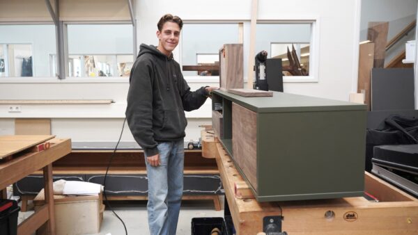 Een student kijkt glimlachend in de camera. Hij is bezig met het maken van een groen dressoir met houten lades.