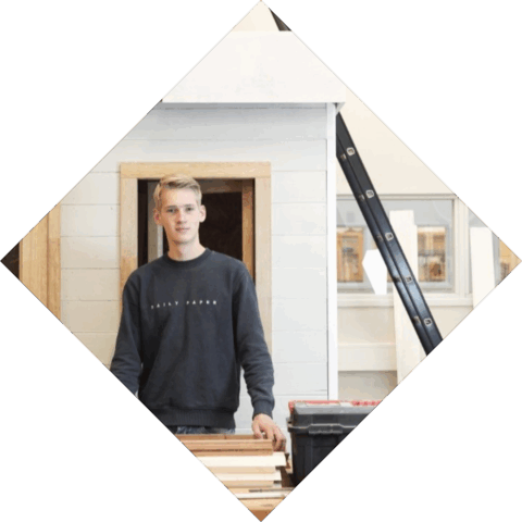 Student staat voor een huisje en leunt op een stapel kozijnen